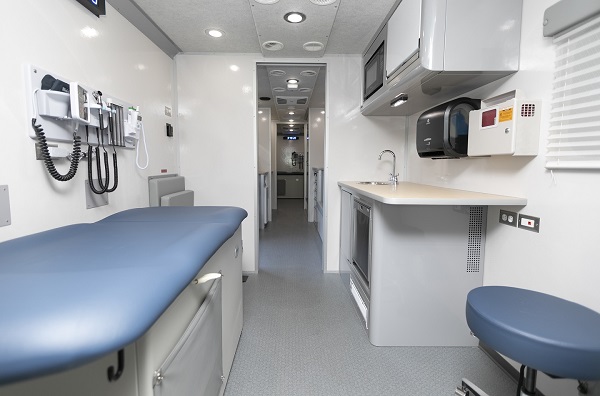 Shows the inside of a mobile clinic. It has a bed on one side, a place to sit, medical equipment on the wall and a wash basin on one the other side.